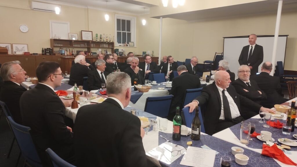Picture of lodge members sitting at the tables during the festive board, with David Mowl standing next to a video screen.