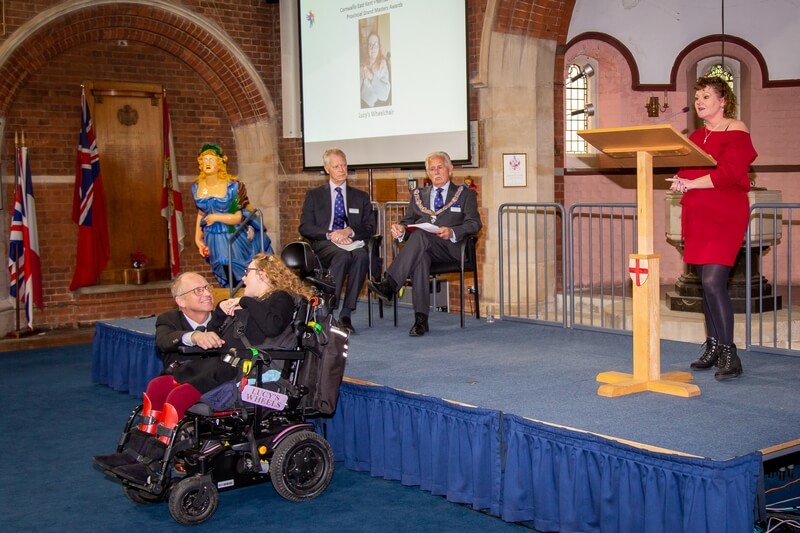 Lady talking on the stage with her daughter in a wheelchair below, talks about Lucys wheels, a family who were helped by the Cornwallis Charity to purchase an electric wheelchair